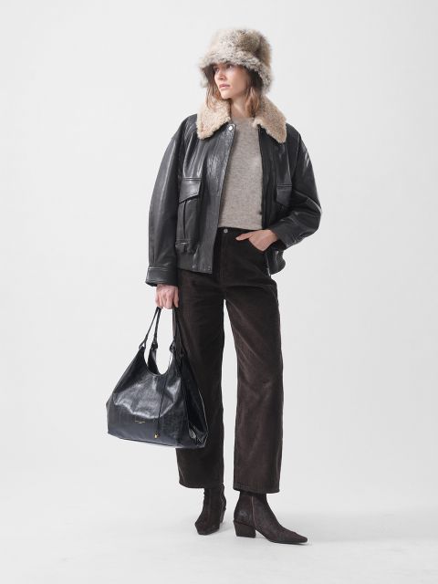 Eine Frau mit einer Pelzmütze, einer schwarzen Lederjacke, einem beigen Pullover, einer braunen Hose und Stiefeln mit Absatz, die eine große schwarze Tasche in der Hand hält und vor einem weißen Hintergrund steht.