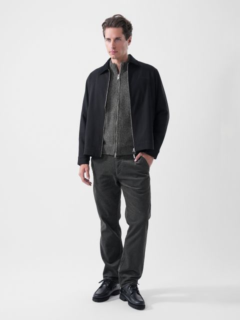 Ein Mann steht vor einem einfarbigen Hintergrund, trägt eine schwarze Jacke, einen grauen Pullover mit Reißverschluss, eine dunkle Hose und schwarze Schuhe und blickt leicht zur Seite.