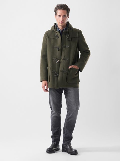 Ein Mann trägt einen olivgrünen Dufflecoat, ein Hemd mit Knopfleiste, dunkelgraue Jeans und schwarze Stiefel.