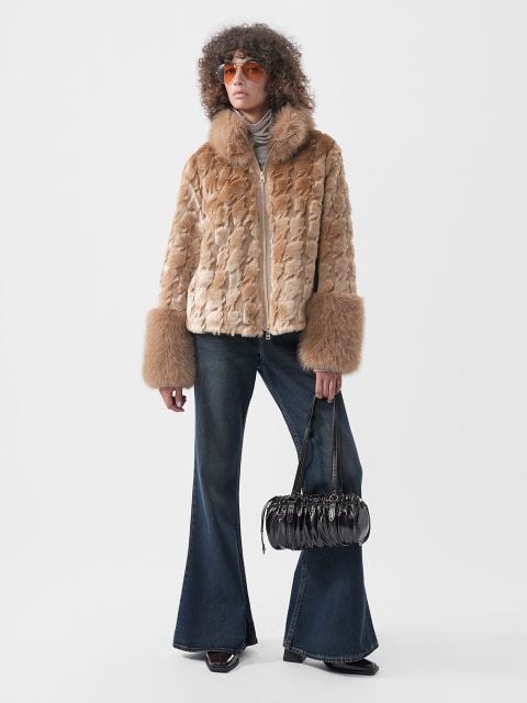 Eine Person, die eine hellbraune Kunstfelljacke mit breiten Ärmelbündchen, eine dunkle Schlagjeans und eine orangefarbene Sonnenbrille trägt und eine schwarze, plissierte Handtasche in der Hand hält.