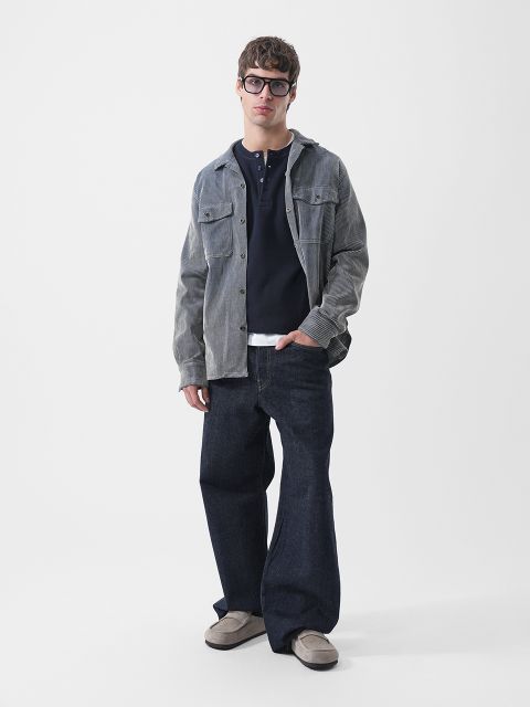 Ein Mann mit einer Brille mit dickem Rahmen, einem aufgeknöpften Cordhemd über einem marineblauen Henley-Shirt, einer dunkelblauen Jeans mit weitem Bein und grauen Loafern.
