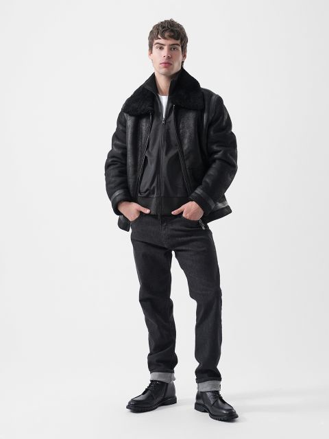 Ein Mann, der eine schwarze, mit Lammfell gefütterte Bomberjacke, schwarze Jeans und schwarze Stiefel trägt.