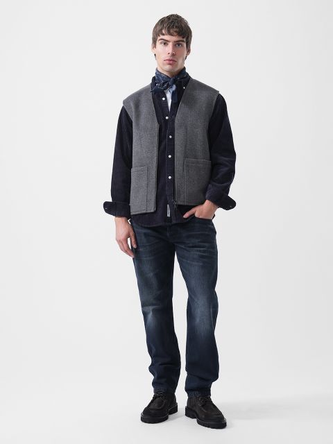 Ein Mann, der eine graue Weste über einem dunklen Hemd mit Knöpfen, ein gemustertes Halstuch, dunkelblaue Jeans und dunkle Stiefel trägt.