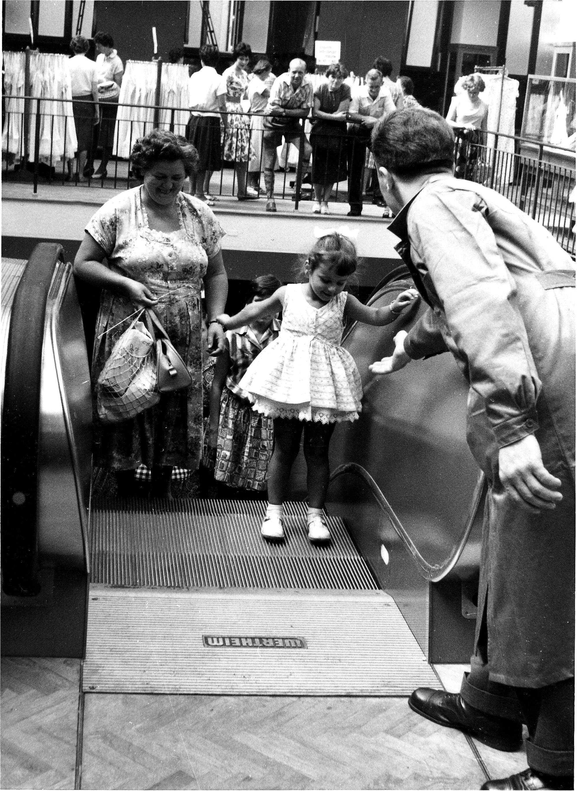 Ein Schwarzweißfoto eines jungen Mädchens in einem weißen Kleid, das mit seiner Mutter eine Rolltreppe betritt. Ein uniformierter Mann hilft ihnen. Im Hintergrund ist eine Menschenmenge in der Nähe von Kleiderständern zu sehen.