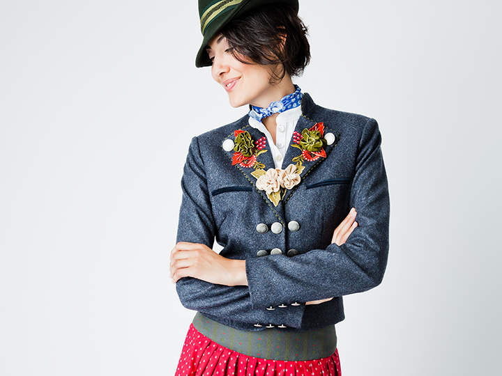 Eine Person in traditioneller Kleidung, bestehend aus einer dunkelblauen Jacke mit Blumenstickereien, einem grünen Hut und einem roten gepunkteten Rock, steht mit verschränkten Armen vor einem einfarbigen Hintergrund.