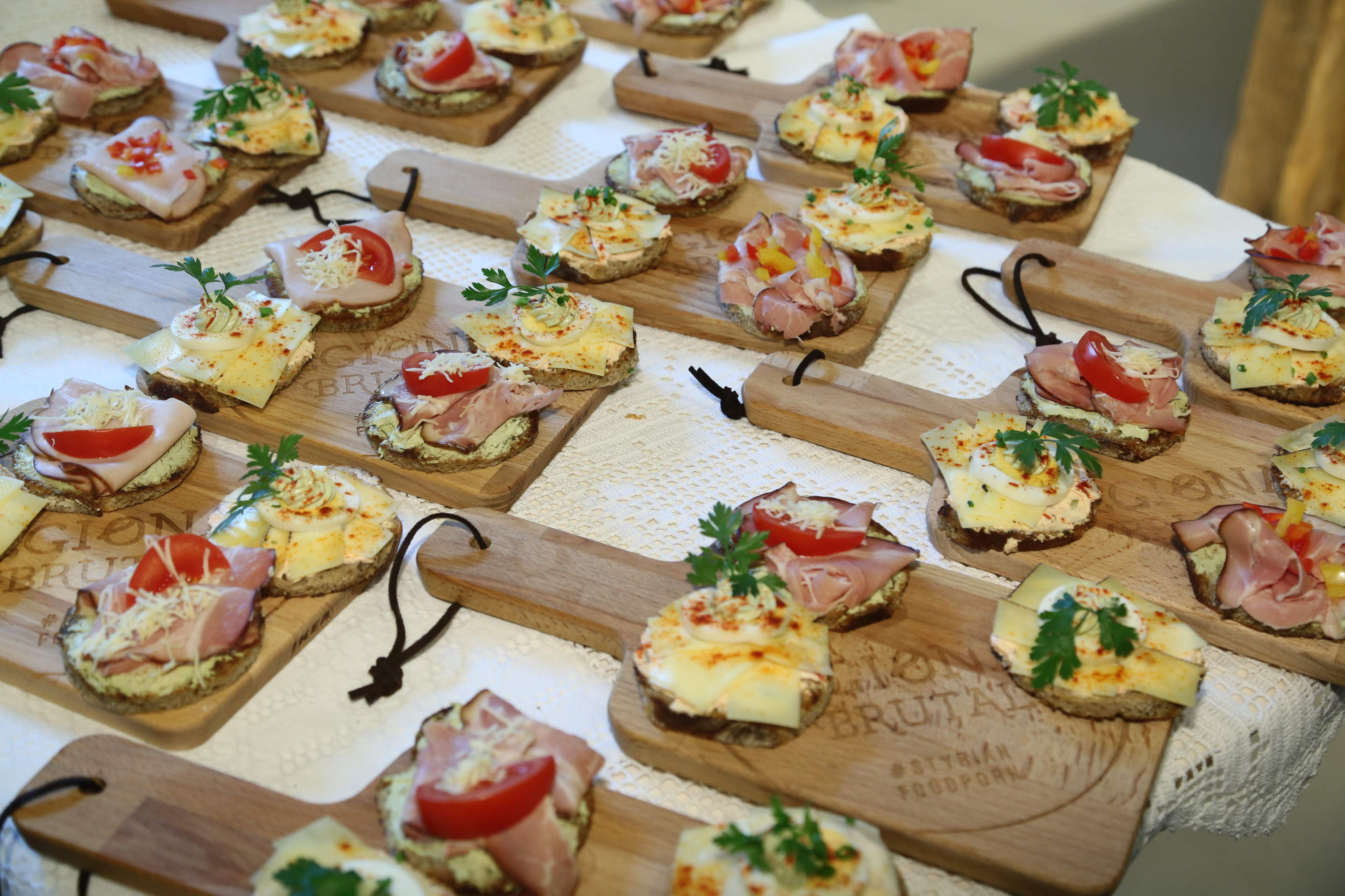 Eine Auswahl verschiedener belegter Brötchen auf Holzbrettern, mit unterschiedlichen Belägen wie Schinken, Käse, Tomatenscheiben, Kräutern und geschnittenem Gemüse, angerichtet auf einer weißen Tischdecke.