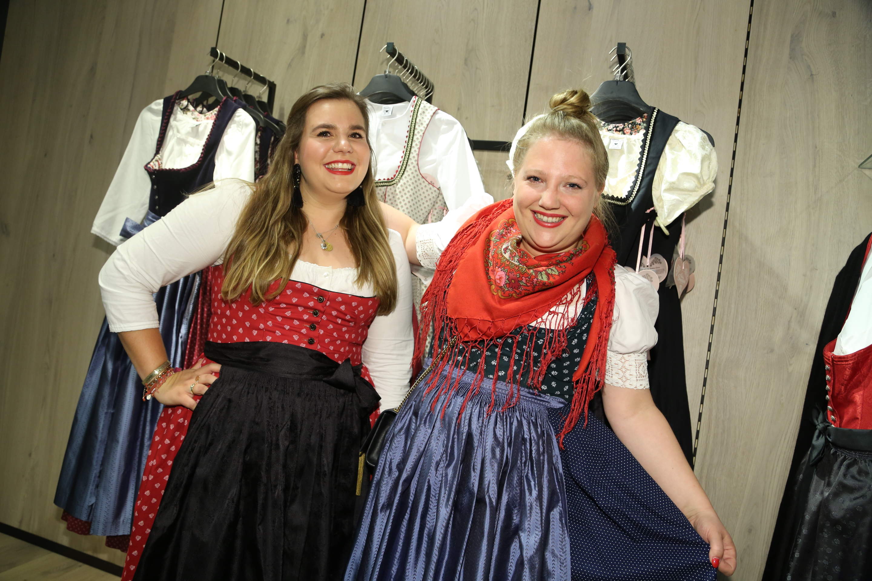 Zwei lächelnde Frauen tragen traditionelle Kleider in einem Holzraum. Die Kleider sind in Rot und Blau gehalten. Hinter ihnen hängen Kleider auf Kleiderbügeln.