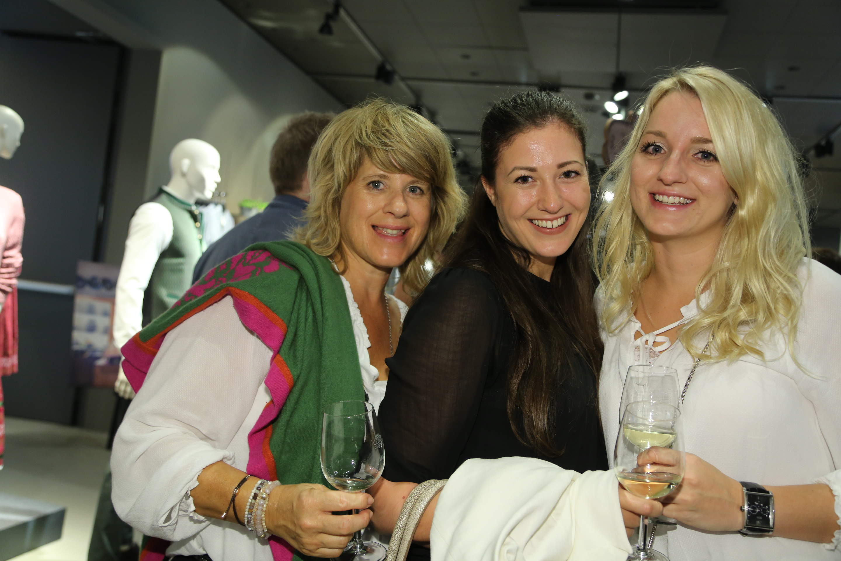 Drei Frauen lächeln und halten Brillen bei einer Indoor-Veranstaltung. Sie tragen lässige, elegante Kleidung. Im Hintergrund sind Schaufensterpuppen in traditioneller Kleidung zu sehen.