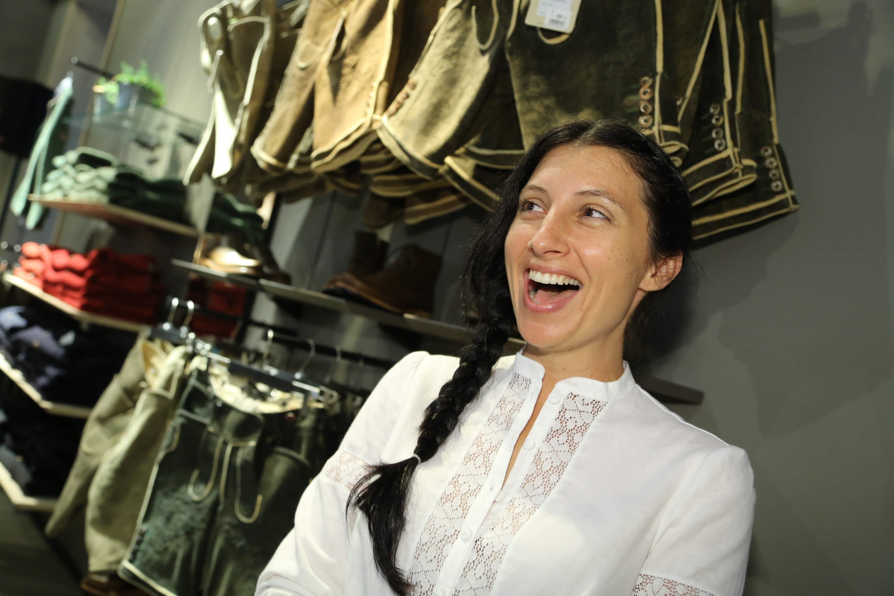 Eine Frau mit langen geflochtenen Haaren und einer weißen Bluse lächelt fröhlich in einem Bekleidungsgeschäft. Im Hintergrund sind verschiedene traditionelle Kleidungsstücke ausgestellt.