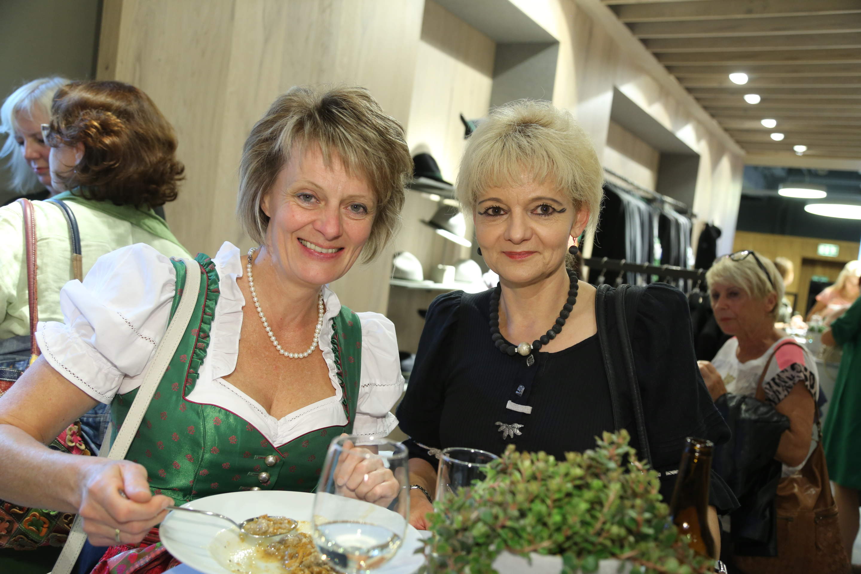 Zwei Frauen lächeln bei einem geselligen Beisammensein. Die Frau links trägt ein traditionelles Dirndlkleid mit einer Perlenkette und hält einen Teller. Die Frau rechts trägt ein schwarzes Outfit mit einer Halskette. Im Hintergrund sind Menschen und Kleiderständer zu sehen.