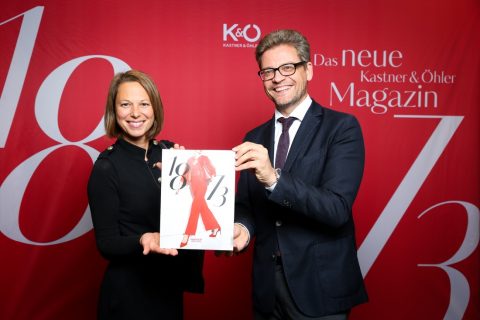 Zwei Menschen lächeln und halten ein Magazin mit einem roten Cover, auf dem eine Modeillustration abgebildet ist. Sie stehen vor einem roten Hintergrund, auf dem der Text „Das neue Kastner & Öhler Magazin“ mit dekorativen Zahlen zu sehen ist.