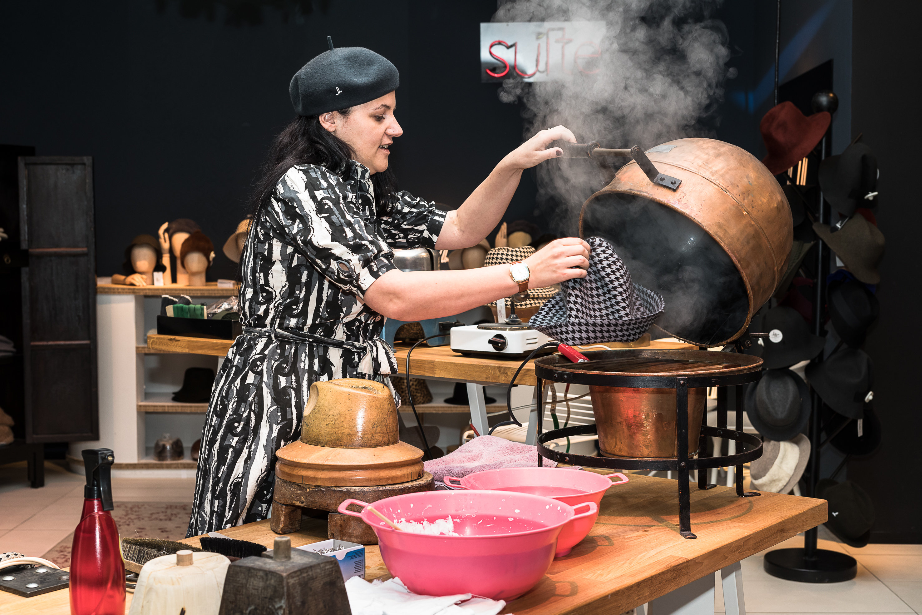 Eine Frau mit Baskenmütze dampft einen Hut über einem großen Kupfertopf. Sie steht an einem Tisch mit verschiedenen Hutmacherwerkzeugen und rosa Schalen. Im Hintergrund sind Regale mit Hüten zu sehen.