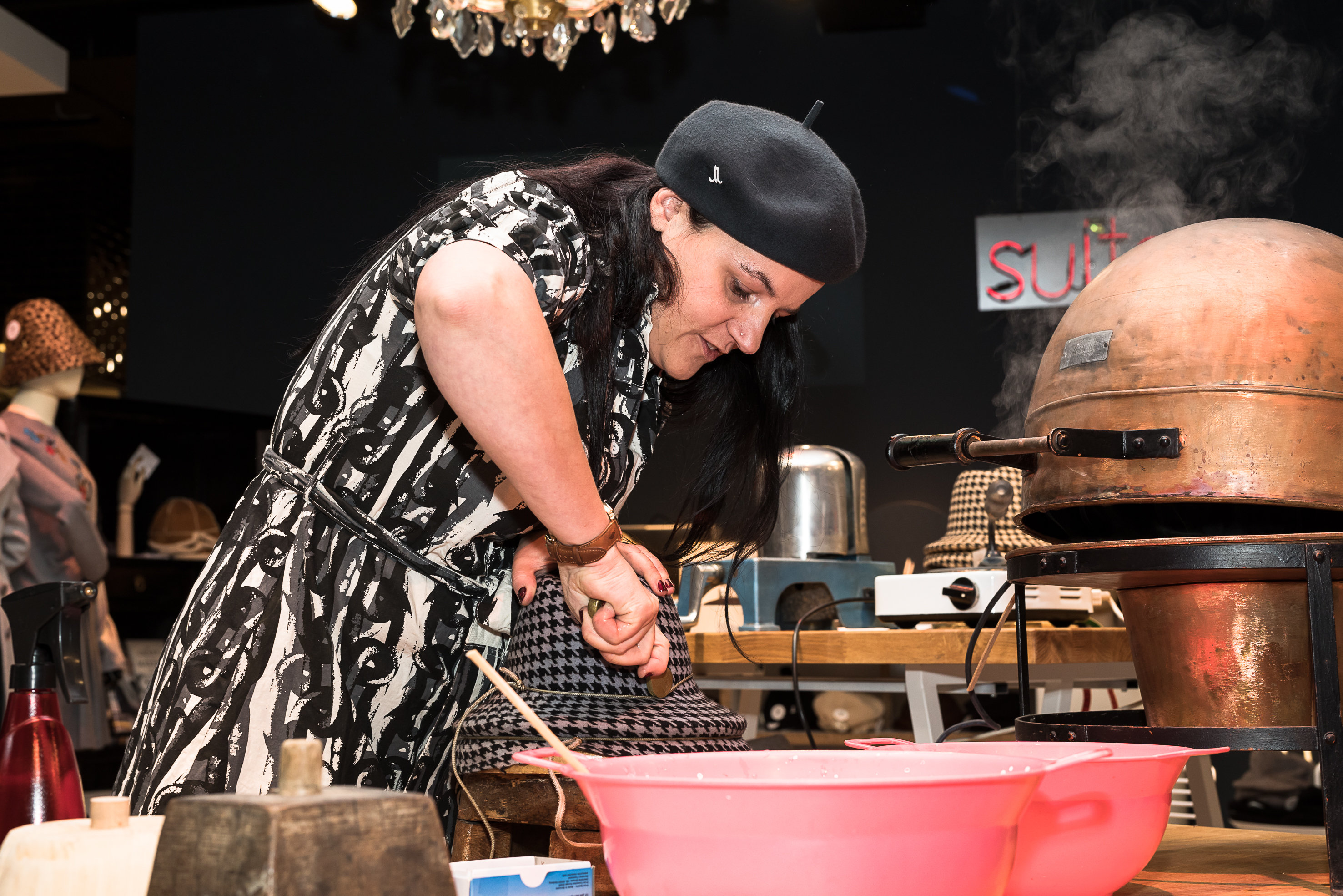 Eine Frau mit schwarzem Barett und gemustertem Kleid bereitet konzentriert Teig an einem Tisch mit Backutensilien zu. Auf dem Tisch stehen eine rosa Rührschüssel und ein großer, kuppelförmiger Ofen. Aus dem Ofen steigt Dampf auf.