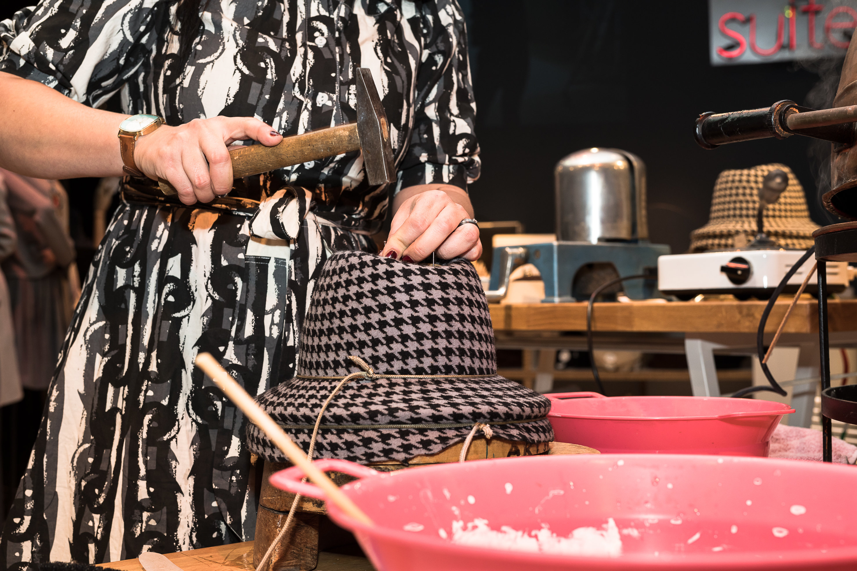 Eine Person in einem gemusterten Kleid bearbeitet mit einem Hammer einen Hut mit Hahnentrittmuster. Die Szene umfasst einen Tisch mit rosa Schüsseln und verschiedenen Werkzeugen, was darauf schließen lässt, dass gerade ein Hut gebastelt oder genäht wird.