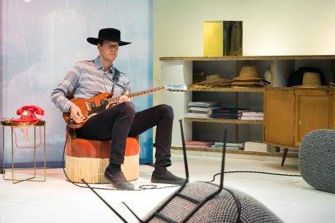 Ein Mann in einem gemusterten Hemd und einem schwarzen Hut sitzt auf einer Ottomane und spielt elektrische Gitarre. In der Nähe steht ein altmodisches rotes Telefon mit Wählscheibe auf einem Ständer. Im Hintergrund sind Regale mit Hüten und Büchern zu sehen. Im Vordergrund steht ein umgekippter Stuhl.