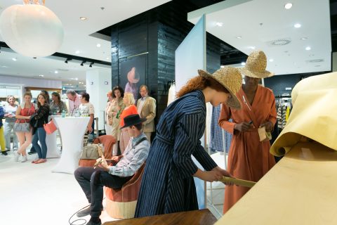 In einem modernen, stilvollen Ausstellungsraum haben sich Menschen in modischer Kleidung versammelt. Zwei Personen mit Strohhüten prüfen an einem Tisch Materialien, während andere zuschauen und sich unterhalten. Eine Person, die auf einem Stuhl sitzt, scheint mit einem Notizblock beschäftigt zu sein.