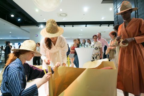 In einem gut beleuchteten Raum verpacken Menschen mit Hüten Gegenstände auf kreative Weise mit Goldpapier. Andere stehen neben einem Cocktailtisch mit Getränken und beobachten das Treiben. Die Atmosphäre ist lebhaft und künstlerisch.