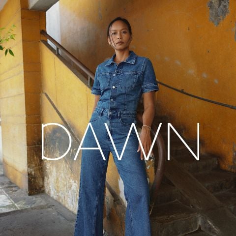 Eine Frau in Jeanskleidung steht selbstbewusst auf einer alten Treppe mit gelben Wänden. Das Wort "DAWN" ist in großen weißen Buchstaben überlagert.