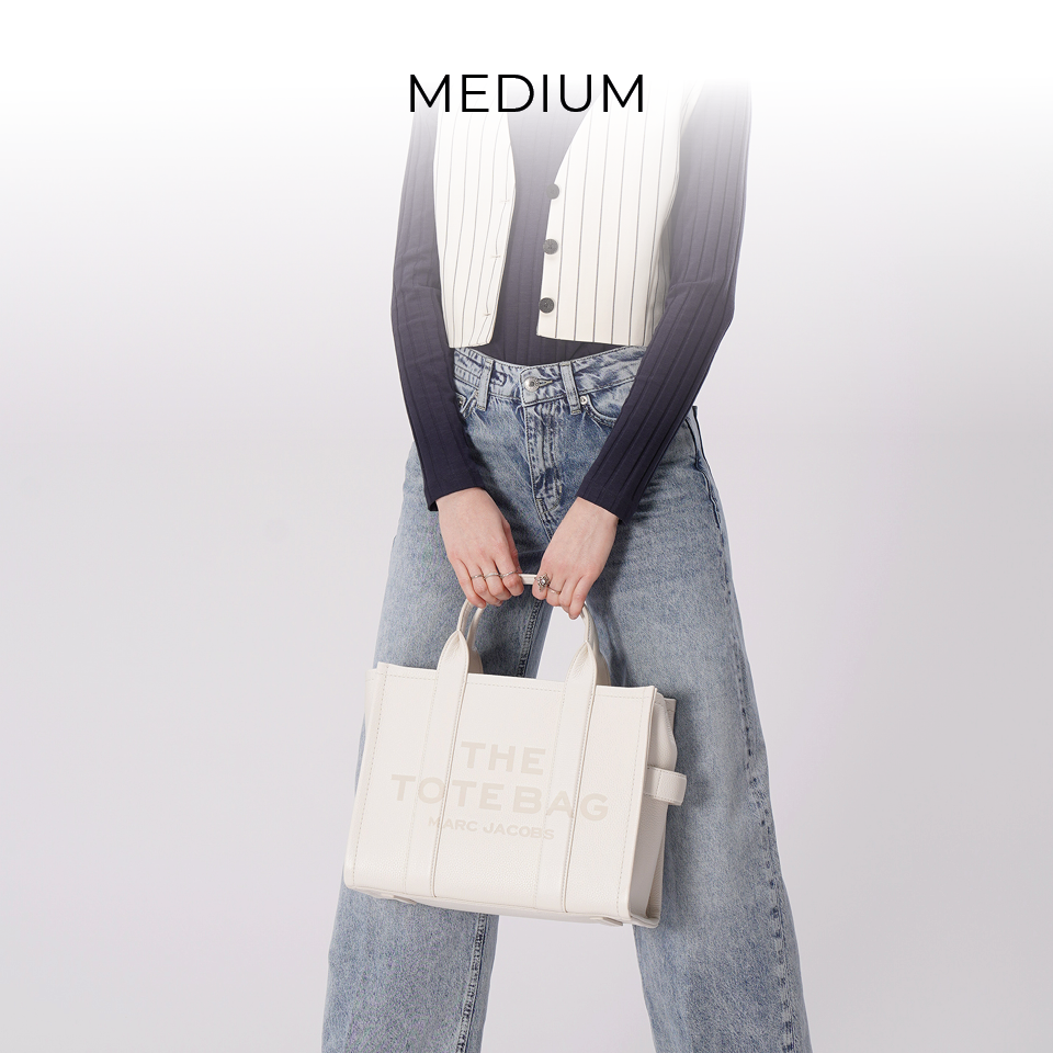 Person hält eine weiße Einkaufstasche mit dem Text „THE TOTE BAG“; trägt ein schwarzes Langarmshirt, eine weiße Weste und locker sitzende Jeans. Darüber steht das Wort „MEDIUM“, das die Größe der Tasche angibt.