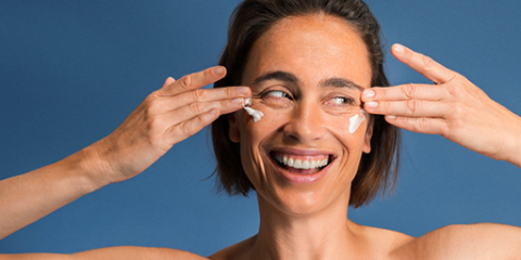 Eine Frau mit kurzen braunen Haaren lächelt, während sie vor einem blauen Hintergrund Creme unter ihren Augen aufträgt. Sie verwendet Zeige- und Mittelfinger, um die Creme sanft zu verteilen.