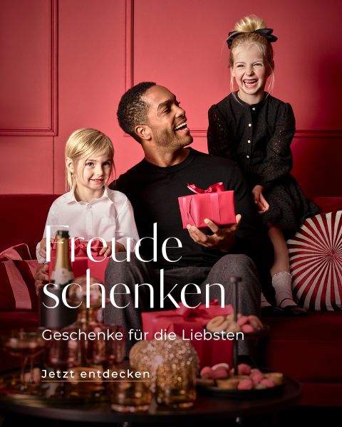 Ein Mann sitzt mit zwei lächelnden Kindern auf einer Couch in einem festlichen, in Rot gehaltenen Raum. Er hält ein verpacktes Geschenk in der Hand und verschiedene Weihnachtsdekorationen sind zu sehen. Der deutsche Text lautet: „Freude schenken. Geschenke für die Liebsten. Jetzt entdecken.“