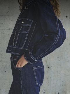 Eine Frau mit weißer Mütze, weißem T-Shirt und blauen Jeans posiert mit ausgestrecktem Arm in einem modernen, weißen Raum mit Blumen darüber.