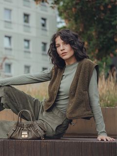 Eine Frau mit gewelltem dunklem Haar sitzt im Freien auf einer erhöhten Fläche und trägt ein grünes, langärmeliges Rippenoberteil, eine olivfarbene Weste und eine passende Hose. Sie hat eine kleine olivgrüne Handtasche neben sich und ein Wohnhaus im Hintergrund.