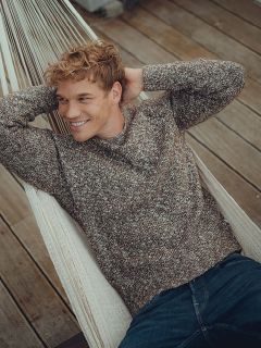 Eine lächelnde Person mit lockigem Haar, die einen braunen Strickpullover und blaue Jeans trägt, liegt mit den Händen hinter dem Kopf in einer weißen Hängematte auf einem Holzdeck.