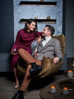 Eine Frau in einem kastanienbraunen Kleid und ein Mann in einem beigen Blazer sitzen zusammen, lächeln und halten Gläser mit Rotwein in einem modernen, stilvollen Wohnzimmer. Eine brennende Kerze steht auf einem Tisch in der Nähe.