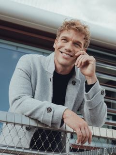 Ein lächelnder Mann mit lockigem blondem Haar lehnt lässig an einem Maschendrahtzaun, trägt einen hellgrauen Mantel über einem schwarzen Hemd und hat ein modernes Gebäude im Hintergrund.