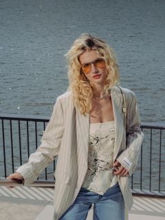 Eine Frau mit lockigem blondem Haar trägt eine Sonnenbrille, einen gestreiften Blazer, ein geblümtes Oberteil, Jeansshorts und weiße Schuhe, während sie an einer Uferpromenade mit Geländer steht.