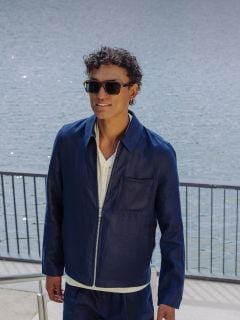 Ein Mann mit Sonnenbrille, marineblauer Jacke und passenden Shorts geht eine Freitreppe an einem Gewässer hinauf, hält sich an einem Geländer fest und lächelt.