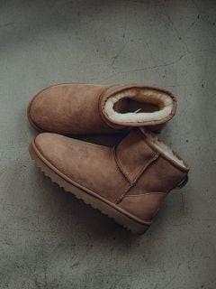 Ein Paar braune, mit Shearling gefütterte Wildlederstiefel stehen auf einem grauen, strukturierten Boden, wobei ein Stiefel den anderen leicht überlappt.