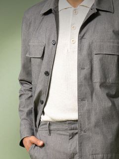 Eine Person, die eine hellgraue, geknöpfte Jacke und eine dazu passende Hose über einem strukturierten weißen Poloshirt trägt, steht mit einer Hand in der Tasche vor einem gedämpften grünen Hintergrund.