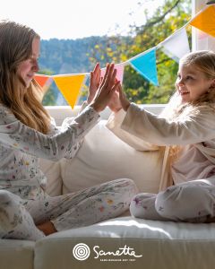 Zwei junge Mädchen in Schlafanzügen sitzen auf einer weißen Couch, lächeln und spielen ein Klatschspiel. Im Hintergrund hängen bunte dreieckige Wimpel, und hinter ihnen fällt das Sonnenlicht durch ein Fenster.