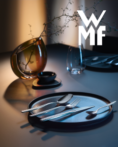 Elegant gedeckter Tisch mit Silberbesteck auf einem schwarzen Teller, einer Glasvase mit Zweigen und dem WMF-Logo im Hintergrund.