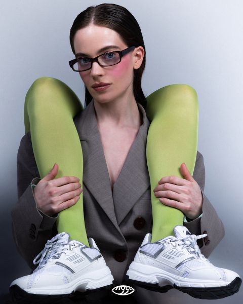 Eine Frau in einem grauen Blazer posiert mit einer hellgrünen Strumpfhose, die sie sich über die Schultern zieht, und weißen Turnschuhen und blickt selbstbewusst in die Kamera.