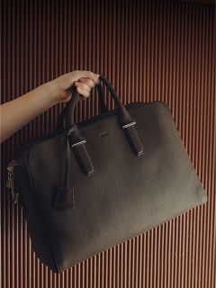 Eine Hand hält eine große, schwarze Ledermappe mit zwei Griffen vor einem braunen, geriffelten Hintergrund.