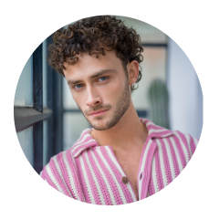 Mann mit lockigen braunen Haaren und blauen Augen, der ein rosa-weiß gestreiftes Hemd trägt und in einem Innenraum an einem Fenster in die Kamera schaut.