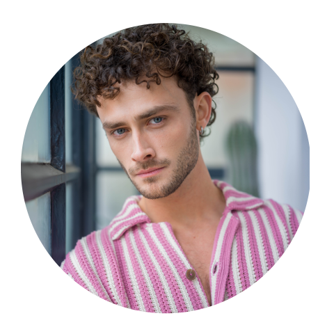 Mann mit lockigen braunen Haaren und blauen Augen, der ein rosa-weiß gestreiftes Hemd trägt und in einem Innenraum an einem Fenster in die Kamera schaut.