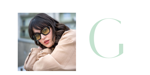 Frau mit dunklem Haar und Sonnenbrille, die einen beigen Mantel trägt und sich auf die Arme stützt, neben einem großen, hellgrünen Buchstaben "G" auf hellem Grund.