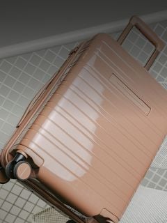 Ein glänzender, hellbrauner Hartschalenkoffer mit Rollen und einem verlängerten Griff steht auf einer gefliesten Fläche.