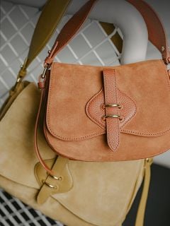 Zwei Handtaschen aus Wildleder, eine hellbraun und eine beige, mit Lederriemen und goldenen Verschlüssen, die auf einem Hintergrund aus weißem Metallgitter hängen.