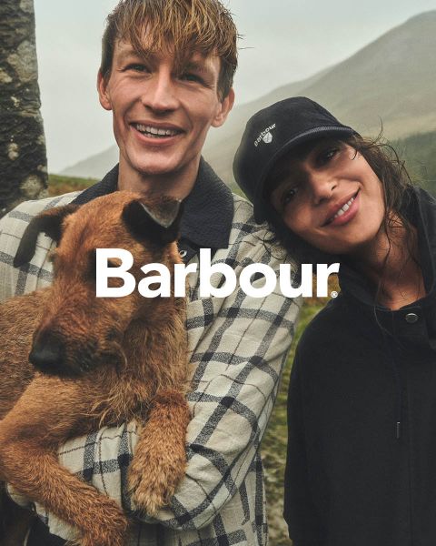 Ein lächelnder Mann, der einen braunen Hund hält, steht neben einer lächelnden Frau mit einer schwarzen Mütze im Freien. Das Wort "Barbour" ist auf dem Bild eingeblendet.