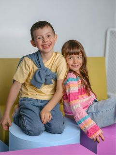 Zwei lächelnde Kinder sitzen auf bunten Schaumstoffblöcken, der Junge in einem gelben Hemd und Jeans, das Mädchen in einem gestreiften Pullover und Jeans, und schauen in die Kamera.