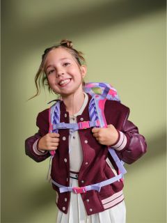 Ein lächelndes junges Mädchen in einer kastanienbraunen Jacke und einem weißen Kleid, das einen bunten Rucksack trägt, steht vor einem grünen Hintergrund.
