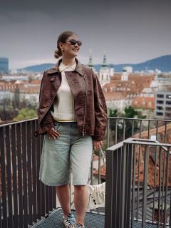 Frau mit Sonnenbrille und brauner Jacke steht auf einem Balkon mit Blick auf eine Stadtlandschaft mit roten Dächern und Kirchtürmen an einem bewölkten Tag.