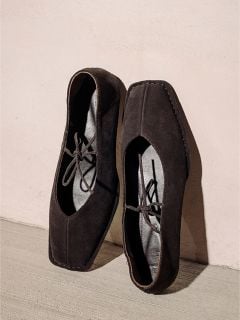 Ein Paar schwarze, quadratische Wildlederschuhe mit Schnürsenkeln, die vor einer hellen Wand und einem hellen Boden stehen.