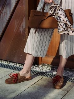 Person, die eine gestreifte Hose mit weitem Bein und braune Slipper mit roten Schnürsenkeln trägt und eine braune Handtasche in der Hand hält, an deren Griff ein Schal mit Leopardenmuster gebunden ist.