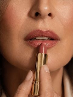 Nahaufnahme einer Frau, die mit einer goldenen Lippenstifttube lila Lippenstift auf ihre Lippen aufträgt.